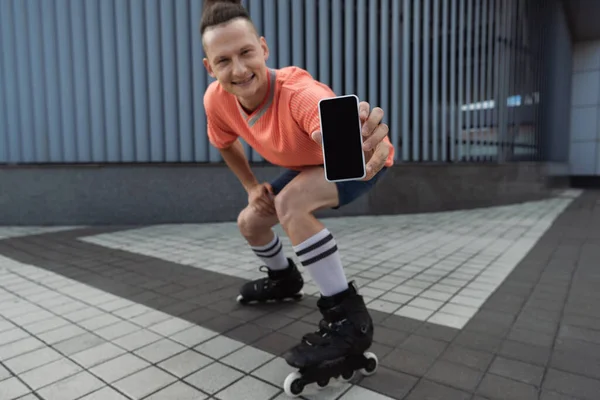 Patinador borroso sonriendo y sosteniendo el teléfono inteligente con pantalla en blanco en la calle - foto de stock