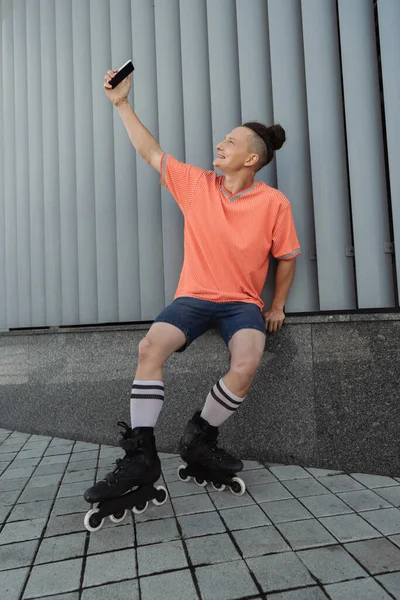 Vista lateral do homem sorridente em patins levando selfie no smartphone perto do prédio ao ar livre — Fotografia de Stock