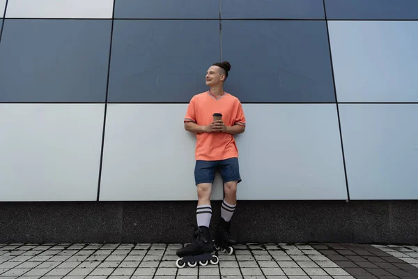 Позитивный человек в роликовых коньках держит бумажную чашку, стоя возле здания на открытом воздухе — стоковое фото
