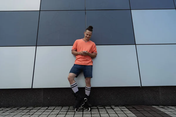 Улыбающийся мужчина в повседневной одежде и роликовых коньках с помощью смартфона возле здания на открытом воздухе — стоковое фото