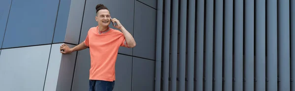Улыбающийся мужчина в футболке разговаривает по мобильному телефону возле здания на открытом воздухе, баннер — стоковое фото
