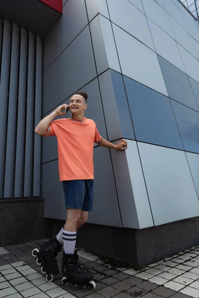 Patinador de rolo alegre falando no smartphone e olhando para longe na rua urbana — Fotografia de Stock
