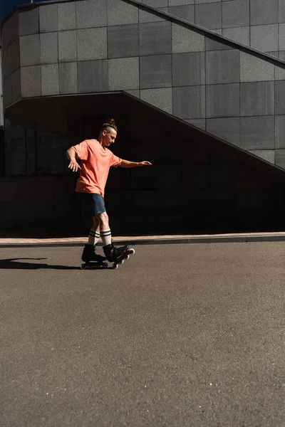 Вид сбоку на роликового конька в шортах и футболке, катающегося на асфальте возле здания на открытом воздухе — стоковое фото