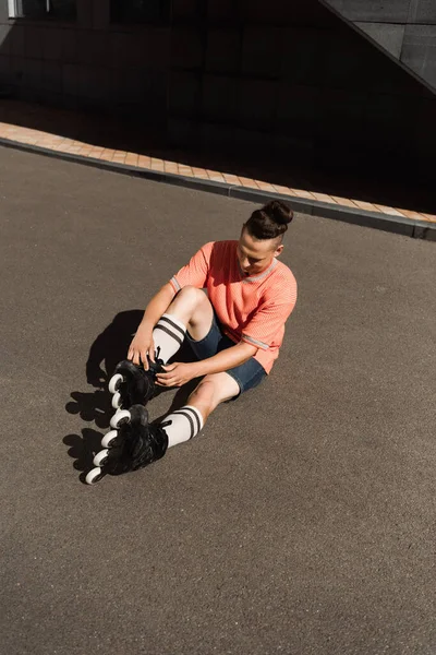 Visão de alto ângulo do jovem vestindo lâminas de rolo enquanto sentado no asfalto na rua urbana — Fotografia de Stock