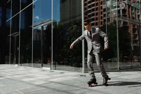 Full length of young businessman in grey suit roller skating near modern building with glass facade — Stock Photo