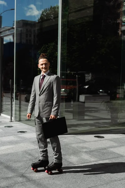 Longitud completa de hombre de negocios sonriente en patines de pie con maletín negro cerca de la fachada de vidrio del edificio moderno - foto de stock