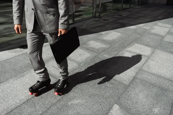 Обрезанный вид бизнесмена в роликовых коньках и сером костюме с черным портфелем на улице в городе — стоковое фото