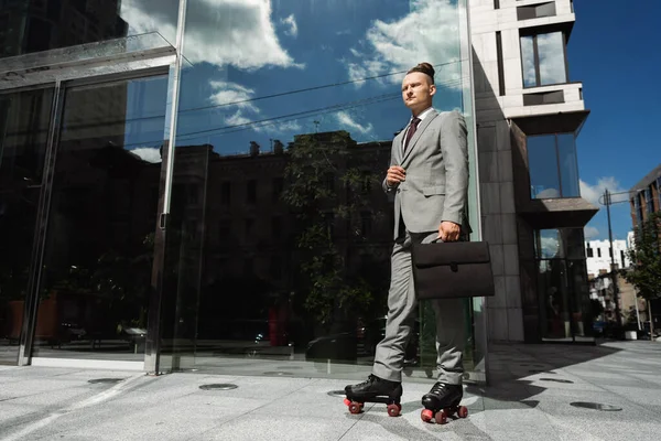Homme d'affaires en costume gris et patins à roulettes debout avec mallette près du bâtiment moderne avec façade en verre — Photo de stock