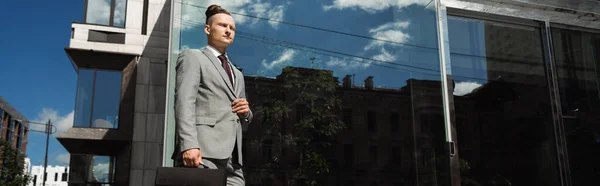 Hombre de negocios joven y de moda en traje gris mirando hacia otro lado en la calle cerca de un edificio moderno con fachada de vidrio, pancarta - foto de stock