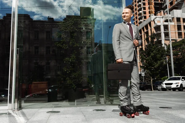 Улыбающийся бизнесмен в роликовых коньках, держащий черный портфель и смотрящий в сторону на городской улице — стоковое фото