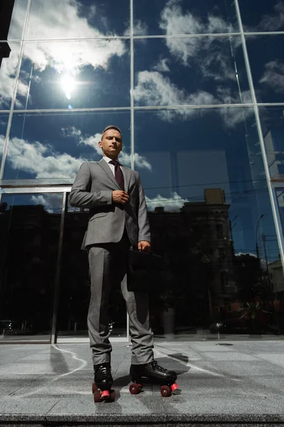 Low angle view of confident businessman in grey suit and roller skates near contemporary building with glass facade — Stock Photo
