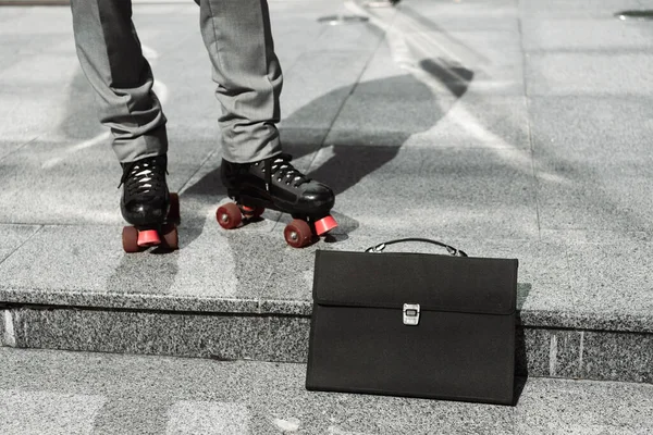 Черный портфель рядом с обрезанным бизнесменом в роликовых коньках на тротуаре городской улицы — стоковое фото