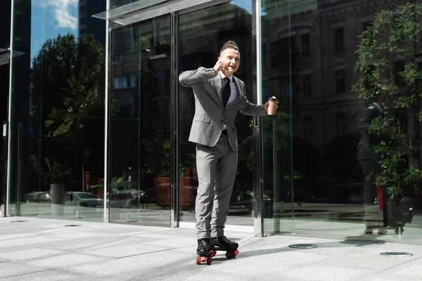 Улыбающийся бизнесмен в роликовых коньках держит выпивку на вынос и разговаривает на смартфоне рядом со стеклянным фасадом современного здания — стоковое фото