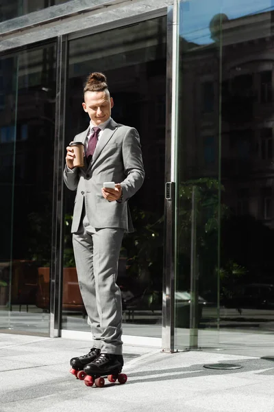 Бізнесмен у костюмі та роликових ковзанах, що тримає каву, щоб піти та обмінюватися повідомленнями на смартфоні біля скляної будівлі — стокове фото