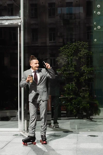 Amazed businessman in roller skates looking away while standing with smartphone and paper cup near glass building — Stock Photo