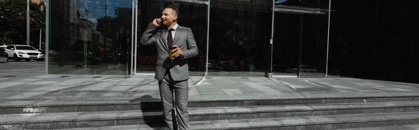 Elegante hombre de negocios en traje gris celebración de bebidas para llevar y hablar en el teléfono celular en la calle de la ciudad, pancarta - foto de stock