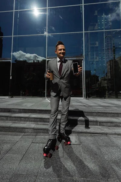 Усміхнений чоловік у костюмі та роликових ковзанах тримає паперову чашку та смартфон із порожнім екраном біля сучасної будівлі — стокове фото