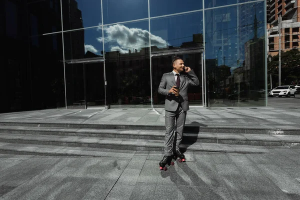 Joven hombre de negocios en patines de ruedas hablando en smartphone y sosteniendo taza de papel cerca del edificio con fachada de vidrio - foto de stock