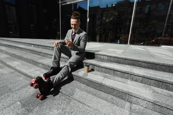 Homme d'affaires positif en patins à roulettes bavarder sur smartphone tout en étant assis près de tasse en papier sur les escaliers — Photo de stock