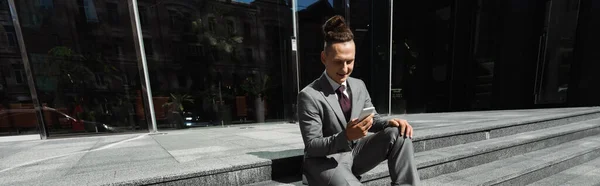 Молодой и позитивный бизнесмен в сером костюме разговаривает на смартфоне, сидя на лестнице на улице, баннер — стоковое фото