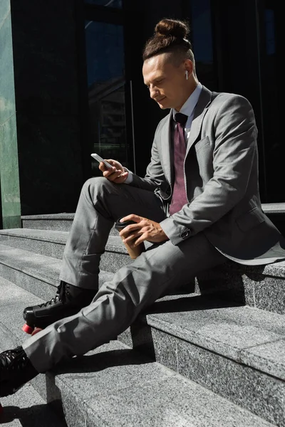 Молодий бізнесмен в роликових ковзанах тримає паперову чашку, сидячи на сходах і обмін повідомленнями на смартфоні — стокове фото