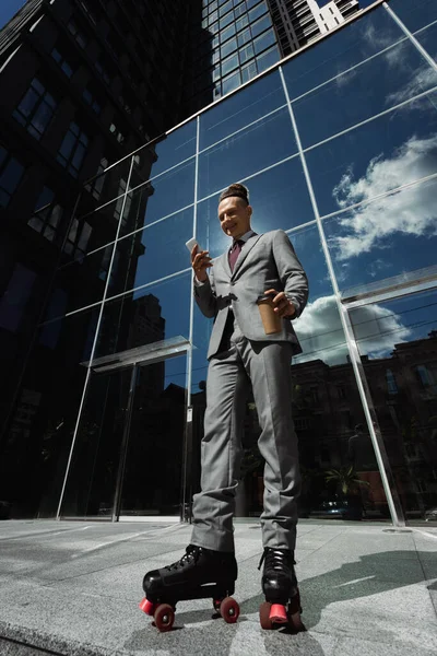 Низький кут зору щасливого ролика в костюмі, що тримає смартфон і виймає напій біля сучасної будівлі зі скляним фасадом — стокове фото