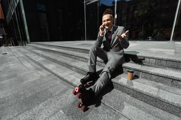 Patinador de rolo sorridente em desgaste formal falando no celular enquanto sentado perto de café para ir em escadas ao ar livre — Fotografia de Stock