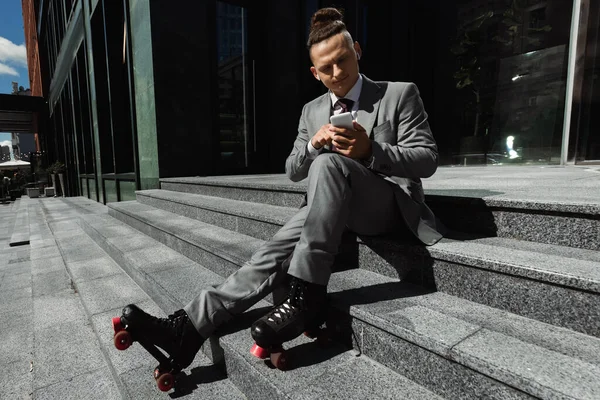 Uomo d'affari in pattini a rotelle e auricolari wireless chatta sul telefono cellulare mentre è seduto sulle scale all'aperto — Foto stock