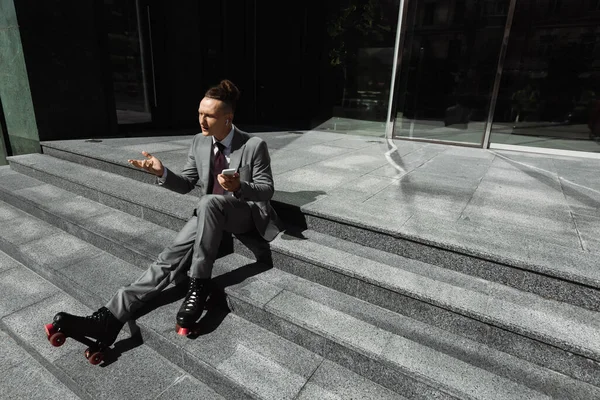 Geschäftsmann in Anzug und Rollschuhen sitzt mit Handy auf der Treppe und zeigt achselzuckende Geste — Stockfoto