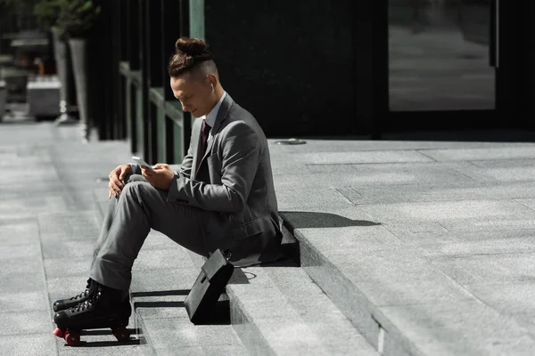 Вид сбоку стильный роликовый конькобежчик в костюме обмена сообщениями на смартфоне, сидя на лестнице рядом с портфелем — стоковое фото
