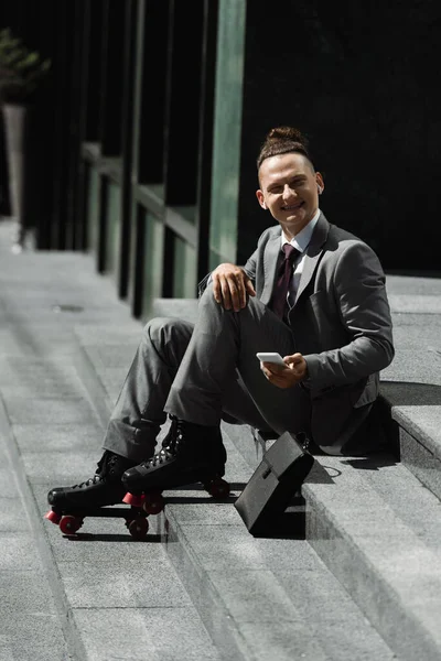Cheerful businessman in roller skates and wireless earphones holding cellphone and looking at camera while sitting on stairs outdoors — Stock Photo