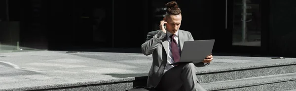 Homem de negócios sério com laptop ajustando fone de ouvido enquanto sentado em escadas na rua, banner — Fotografia de Stock