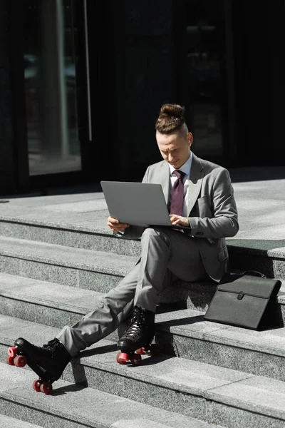 Повна довжина бізнесмена в роликових ковзанах, сидячи з ноутбуком біля портфеля на сходах міської вулиці — стокове фото
