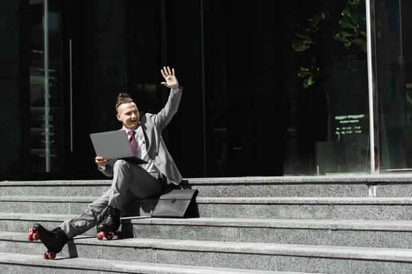 Homme souriant en costume et patins à roulettes assis sur les escaliers avec ordinateur portable et agitant la main tout en regardant loin — Photo de stock