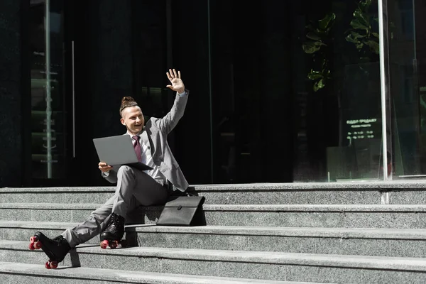 Sonriente hombre de traje y patines de ruedas sentado en las escaleras con el ordenador portátil y la mano agitada mientras mira hacia otro lado - foto de stock