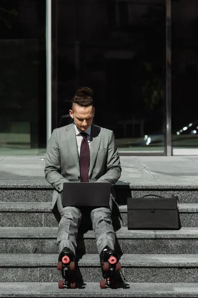 Stylischer Geschäftsmann in formeller Kleidung und Rollschuhen, der auf einem Laptop tippt, während er auf einer Treppe neben der Aktentasche sitzt — Stockfoto