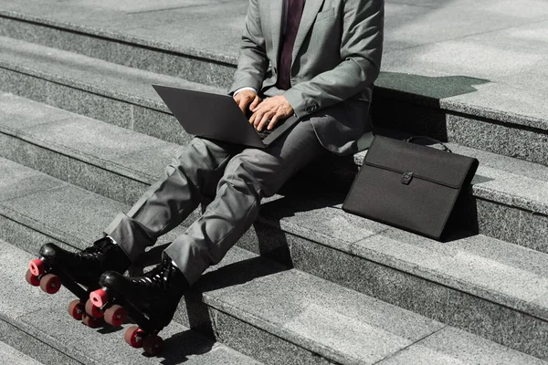 Обрезанный вид бизнесмена в сером костюме и роликовых коньках, сидящего на лестнице рядом с портфелем и печатающего на ноутбуке — стоковое фото