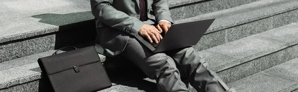 Vista parcial del hombre de negocios en traje gris sentado en las escaleras cerca del maletín negro y escribir en el ordenador portátil, pancarta - foto de stock
