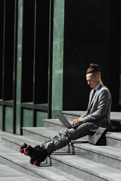 Comprimento total do homem em terno cinza e patins sentados nas escadas perto da pasta e digitando no laptop — Fotografia de Stock