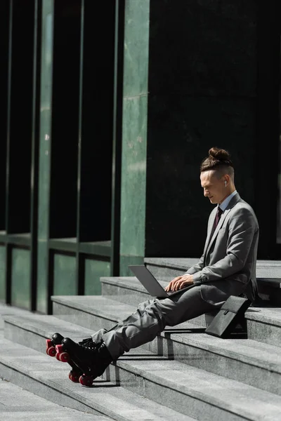 Full length of man in grey suit and roller skates sitting on stairs near briefcase and typing on laptop — Stock Photo