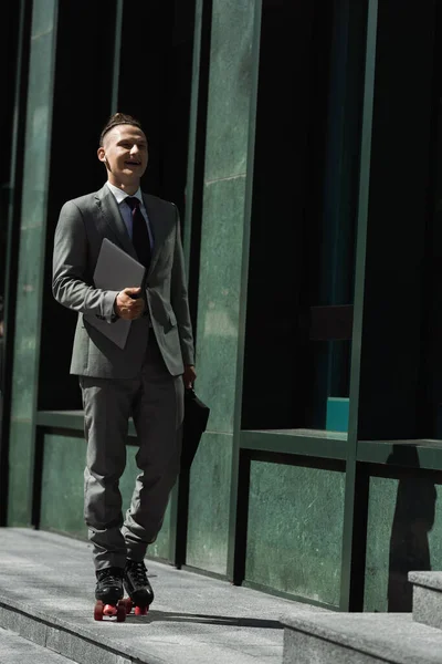 Uomo d'affari sorridente in giacca e cravatta e pattini a rotelle in sella con laptop e valigetta su strada urbana — Foto stock