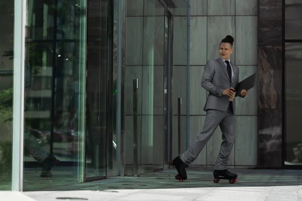 Jeune homme d'affaires en patins à roulettes et costume gris tenant café pour aller et ordinateur portable près du bâtiment avec portes en verre — Photo de stock
