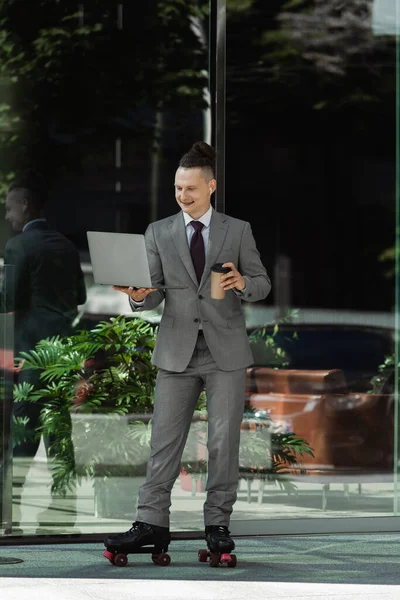 Smiling man in grey suit and roller skates holding coffee to go and looking at laptop near green plant behind glass facade — Stock Photo