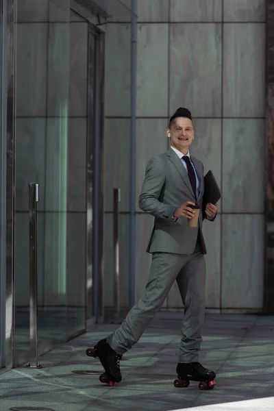 Улыбающийся бизнесмен на роликовых коньках с ноутбуком и бумажной чашкой во время катания из здания со стеклянными дверями — стоковое фото
