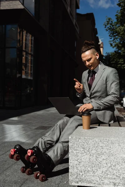 Молодий чоловік в роликових ковзанах і діловому костюмі сидить з ноутбуком біля кави, щоб піти і вказувати пальцем — стокове фото