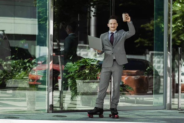Uomo d'affari sorridente in pattini a rotelle in piedi con computer portatile e caffè per andare in mano alzata vicino edificio di vetro — Foto stock