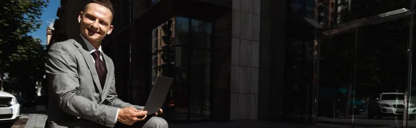 Щасливий бізнесмен у сірому костюмі, сидячи з ноутбуком і дивлячись на камеру на міській вулиці, банер — стокове фото