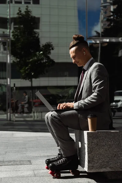 Side view of roller skater in grey suit and wireless earphone typing on laptop near coffee to go on urban street — Stock Photo