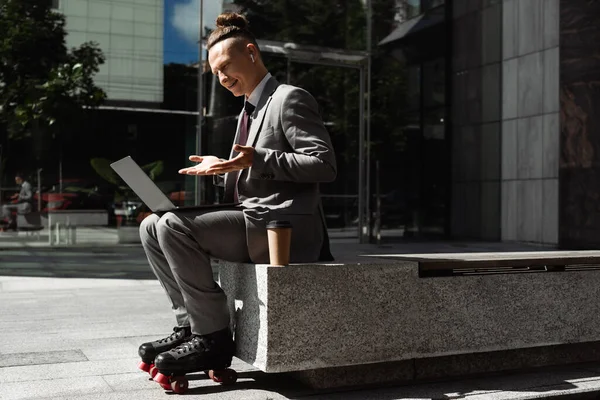 Uomo d'affari sorridente in pattini a rotelle che punta al computer portatile durante videochiamata sulla strada della città — Foto stock