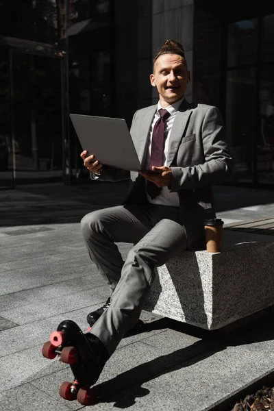 Cheerful businessman in suit and roller skates holding laptop and looking at camera on city street — Stock Photo